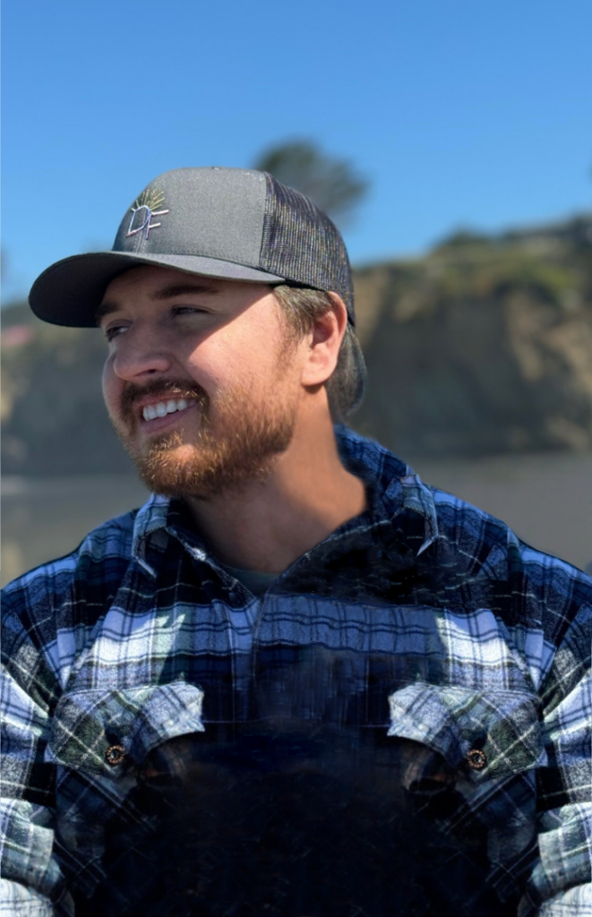 Christian, Reno NV personal trainer and owner of Divine Fitness, smiling at the beach in a flannel jacket and hat under clear blue skies.